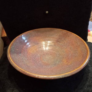 Puede incluir: Un cuenco de cerámica hecho a mano con un diseño ancho y poco profundo. El cuenco presenta una paleta de colores terrosos moteados con tonos de marrón, morado y dorado. El borde tiene una fina banda marrón dorada. Este cuenco decorativo es adecuado para servir o exhibir.