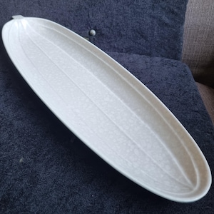 Op de afbeelding: Een langwerpige, ovale serveerschaal in gebroken wit met een textuuroppervlak en verticaal geribbeld ontwerp. Het keramische gerecht is geschikt voor het serveren van eten of het presenteren van decoratieve items. De schaal is ongeveer 46 cm lang.