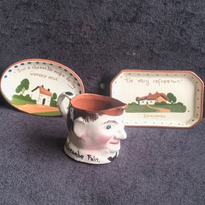 Pode incluir: Um conjunto de itens de cerâmica vintage, incluindo um jarro em forma de rosto masculino e dois pratos pequenos. O jarro tem a inscrição "Widecombe Fair". Os pratos exibem cenas pintadas e texto.