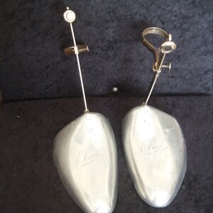 May include: Pair of silver-colored shoe trees with adjustable metal components. Each tree has a curved, shoe-shaped form with the brand name "Clark" embossed on the surface. The trees are designed to maintain the shape of shoes.