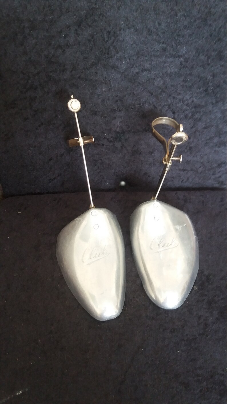 May include: Pair of silver-colored shoe trees with adjustable metal components. Each tree has a curved, shoe-shaped form with the brand name "Clark" embossed on the surface. The trees are designed to maintain the shape of shoes.
