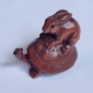 May include: A small, hand-carved brown wooden figurine depicting a rabbit sitting on top of a turtle. The turtle has a detailed, segmented shell and an extended neck, while the rabbit is positioned with its ears back and paws resting on the shell.