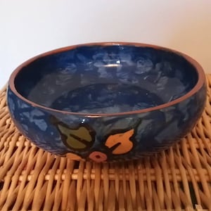 Op de afbeelding: Een handgemaakte keramische kom met een diepblauwe binnenkant en een roodbruine rand. De kom heeft handgeschilderde bloemmotieven in groen, oranje en roze. De kom staat op een geweven rieten oppervlak.