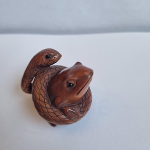 May include: A small, hand-carved brown wooden figurine depicting a snake coiled tightly around a frog. The sculpture features detailed scales on the snake's body and smooth, polished surfaces on the frog, with dark, inlaid eyes on both creatures.