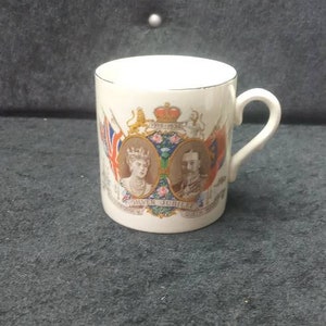 May include: White ceramic mug with a handle, featuring portraits, flags, and text. The mug has a gold rim and is likely a commemorative item. The design includes a crown and the text "Silver Jubilee".