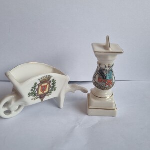Könnte beinhalten: Zwei kleine, dekorative Porzellanfiguren. Eine ist eine weiße Schubkarre mit Wappen. Die andere ist eine weiße Sonnenuhr mit dekorativem Sockel und goldenen Akzenten. Beide haben goldene Verzierungen.