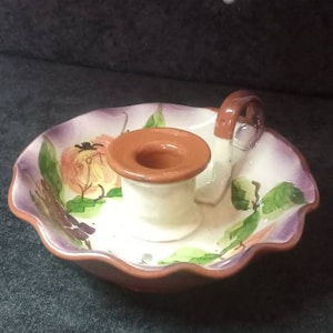 Puede incluir: Un candelero de cerámica con borde festoneado. El soporte tiene una base blanca con borde marrón y un asa marrón. El plato está decorado con flores y hojas pintadas en tonos morado, naranja y verde.
