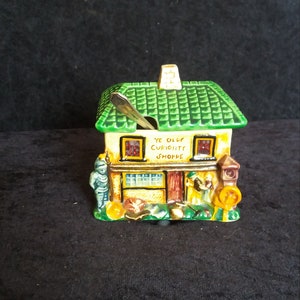 May include: A ceramic house-shaped container with a green roof and the words "Ye Olde Curiosity Shoppe." The container has a small chimney and a silver spoon. Figures of people and objects are painted on the front.