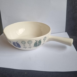 以下が含まれることがあります： クリーム色のセラミック製サービングボウル、持ち手付き。ボウルは青、緑、白の花柄で装飾されています。持ち手はボウルと同じ色です。ボウルは白い表面に置かれています。