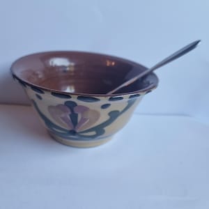 Può includere: Una ciotola in ceramica con interno marrone ed esterno color crema. La ciotola è decorata con motivi floreali nei toni del viola, verde e beige. Un cucchiaio d'argento è all'interno della ciotola. La ciotola ha un aspetto rustico e fatto a mano.