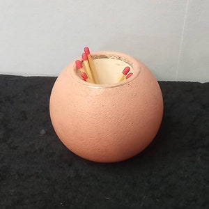 Puede incluir: Un portafósforos esférico de color melocotón con una superficie texturizada. Varios fósforos de madera con puntas rojas son visibles en el interior. El portafósforos está sobre una superficie oscura.