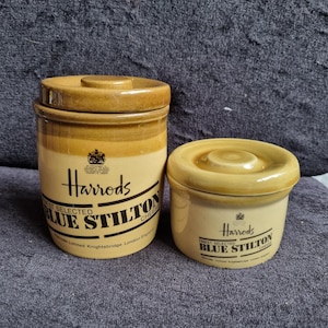 May include: Two ceramic jars with lids. The larger jar is cylindrical, and the smaller one is squat. Both are tan with a cream-colored band and feature the "Harrods" logo and "Blue Stilton Cheese" text. The jars are on a dark blue surface.
