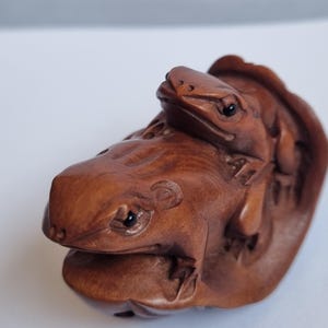 May include: A small, hand-carved wooden figurine featuring two frogs positioned on a curved, leaf-like base. The sculpture is crafted from a medium-brown wood with a smooth, polished finish, highlighting the detailed textures of the frogs' skin.