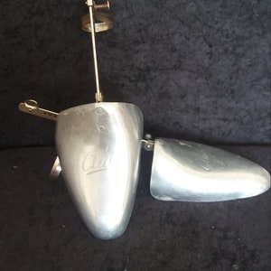 May include: A vintage metal shoe stretcher with a silver finish. The shoe stretcher is open, revealing the interior. The word "Club" is engraved on the main body. A small metal ball sits below the shoe stretcher.