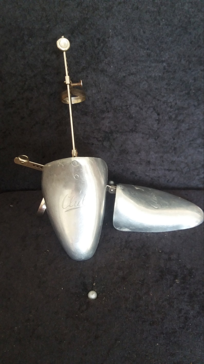 May include: A vintage metal shoe stretcher with a silver finish. The shoe stretcher is open, revealing the interior. The word "Club" is engraved on the main body. A small metal ball sits below the shoe stretcher.