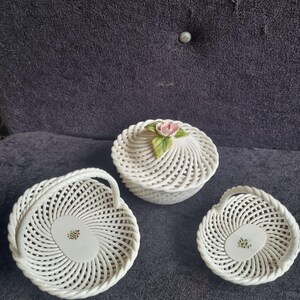 May include: Three white ceramic decorative pieces. A lidded round container with a pink rose and green leaves on top, a small basket with a handle, and a small dish. All pieces have a woven design.