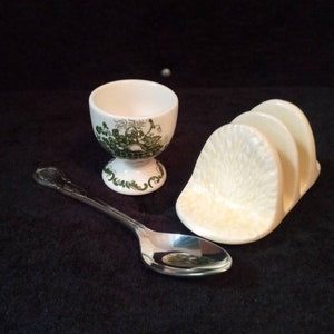 May include: A white egg cup with a green floral design, a silver spoon, and a cream-colored toast rack are arranged on a dark surface. The egg cup has a pedestal base. The spoon has a decorative handle. The toast rack has three slots.