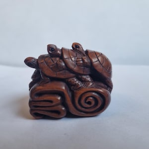 May include: A small, dark brown carved wooden figurine depicting three turtles stacked on top of each other. The turtles are perched upon a base featuring a prominent, deeply carved spiral design and layered, wave-like patterns on the side.