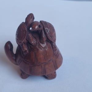 May include: A dark brown carved wooden figurine depicting a large turtle with two smaller turtles resting on its shell. The sculpture features detailed textures on the shells and limbs, with small black inlaid eyes on each of the three turtle figures.