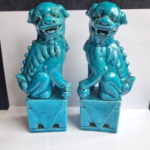 Puede incluir: Dos estatuas de perros Foo de cerámica turquesa. Cada perro está sentado sobre una base cuadrada con recortes decorativos. Los perros tienen detalles, incluyendo bocas abiertas, y son de un vibrante tono azul-verdoso.