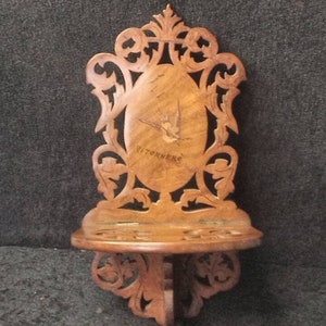 以下が含まれることがあります： 装飾的な木製の壁掛け棚。彫刻が施されたデザインで、中央に鳥のイラストと「Titterne」の文字が彫られています。下部には小さな丸い棚があります。