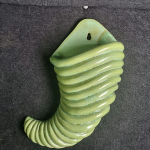May include: A green ceramic wall pocket in the shape of a cornucopia. The wall pocket has a textured, ribbed design and a hole for hanging. The color is a muted, light green with a glossy finish.
