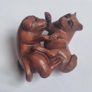 May include: A small, hand-carved brown wooden figurine depicting two animals interacting. The sculpture features smooth, polished surfaces with detailed facial expressions, dark inlaid eyes, and visible paw and tail textures on both figures.