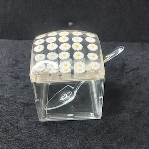 以下が含まれることがあります： 白いヒナギクと黄色の中心で飾られた蓋が付いた、透明なアクリル製の角砂糖入れ。お揃いの透明なスプーンが容器の中にあります。角砂糖入れは正方形で、暗い表面に置かれています。