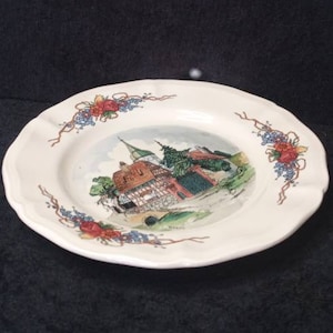 Puede incluir: Plato de cerámica blanca con borde festoneado. El centro presenta una colorida ilustración de una escena de pueblo europeo con una iglesia. Diseños florales y de vid decoran el borde.