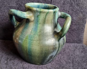 Pode incluir: Um vaso de cerâmica artesanal com duas alças. O vaso apresenta um corpo arredondado e uma abertura larga. O exterior é esmaltado com uma mistura de tons verdes, azuis e castanhos, criando uma aparência artística única. Uma peça decorativa.