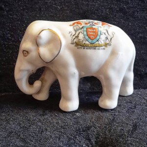 May include: A white ceramic elephant figurine with gold accents. The elephant has a detailed design and a crest on its back with the text "CITY OF HEREFORD A.D. 1645". The figurine is on a dark blue surface.