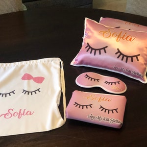 May include: A pink satin pillow with the name "Sofia" and eyelashes printed on it. The pillow is part of a sleep set that includes a matching eye mask and a drawstring bag with the same design. The bag has a pink bow and the text "Sofia" printed on it.