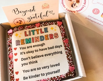 May include: A chocolate brownie with pink and white sprinkles, topped with a white card that reads "Little Reminders" and includes positive affirmations. The brownie is in a white box with a pink and white logo that says "The Happiness Bakery".