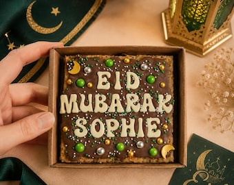 May include: A square chocolate dessert in a cardboard box, decorated with white frosting that spells "EID MUBARAK SOPHIE". The dessert is adorned with green, silver, and gold sprinkles and crescent moon accents. A green ribbon and card with a crescent moon are also visible.
