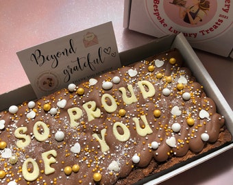 Puede incluir: Un brownie de chocolate con letras de chocolate blanco que deletrean "SO PROUD OF YOU" y chispas doradas y blancas. El brownie está en una caja blanca con una tarjeta que dice "Beyond grateful" y un pequeño gráfico de sobre.