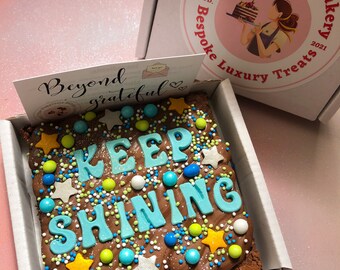 May include: A chocolate brownie topped with chocolate frosting and colorful sprinkles. The brownie has the words "KEEP SHINING" spelled out in blue frosting. The brownie is in a white box with a pink lid. The lid has a logo for "The Happiness Bakery" with a cartoon baker holding a cake.