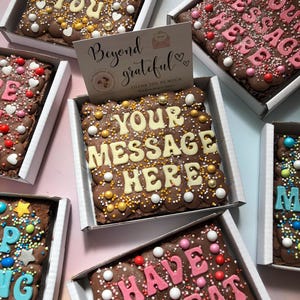 Puede incluir: Brownies de chocolate en cajas blancas, decorados con chispas de colores y mensajes como "Thank You" y "Your Message Here" en glaseado blanco. Una tarjeta dice "Beyond grateful".