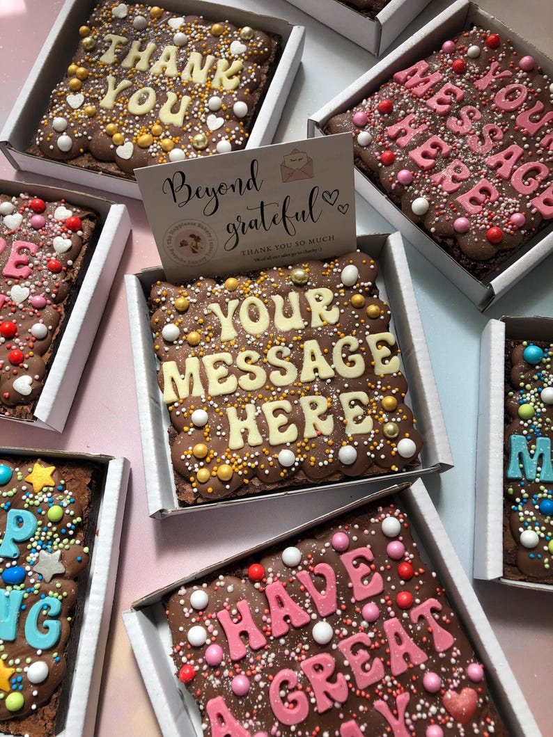 Puede incluir: Brownies de chocolate en cajas blancas, decorados con chispas de colores y mensajes como "Thank You" y "Your Message Here" en glaseado blanco. Una tarjeta dice "Beyond grateful".