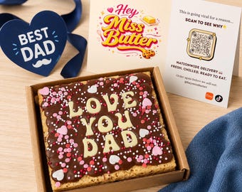 May include: A square chocolate frosted dessert in a cardboard box, decorated with white letters that spell "LOVE YOU DAD". The dessert is surrounded by pink, red, and white sprinkles. A blue heart-shaped decoration with the words "BEST DAD" is also present.