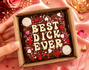 May include: A chocolate brownie in a cardboard box, decorated with white letters spelling "BEST DICK EVER". The brownie is covered in red, pink, and white sprinkles and heart-shaped candies. Two heart-shaped candies are on the top of the brownie, one says "MARRY ME" and the other says "LOVE".
