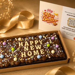 May include: A chocolate brownie in a cardboard box, decorated with colorful sprinkles and pastel-colored candies. The brownie spells out "HAPPY NEW HOME" in white icing. A card with the text "Hey Miss Butter" is visible.