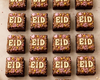 May include: Sixteen square chocolate brownies, each decorated with the word "EID" in white icing. The brownies are topped with pink and gold sprinkles and small pink heart-shaped candies. The arrangement is on a light-colored surface.