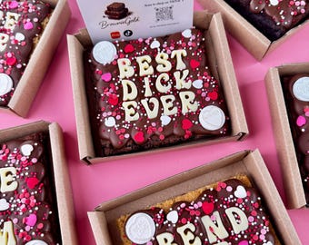 May include: Several chocolate brownies in cardboard boxes, decorated with white icing lettering and colorful sprinkles. The brownies have phrases like "Happy Vals Day" and "Best Dick Ever". A business card is also visible.
