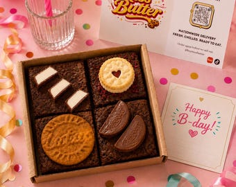 May include: A square cardboard box containing four brownies topped with various cookies and chocolates. One brownie has white chocolate pieces, one has a jam-filled biscuit, one has a Lotus Biscoff cookie, and one has two chocolate segments.