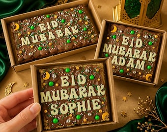 May include: Three chocolate desserts in cardboard boxes, each decorated with the words "EID MUBARAK" in white icing. The desserts are adorned with green, silver, and gold sprinkles, and a crescent moon. One box has the name "Sophie".