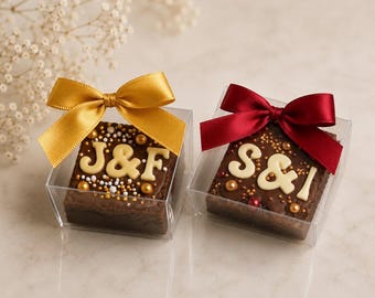 May include: Two square brownies in clear boxes, each topped with initials and decorative sprinkles. One brownie has gold sprinkles and a gold bow, the other has red sprinkles and a burgundy bow. Ideal for wedding favors.