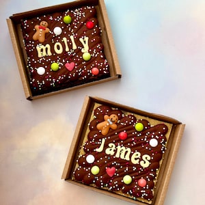 Puede incluir: Dos brownies de chocolate cuadrados en cajas de cart&oacute;n. Cada brownie est&aacute; decorado con glaseado de chocolate, chispas y caramelos de colores. Uno dice "Molly" y el otro "James" en glaseado blanco, con hombres de jengibre.