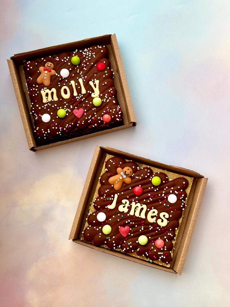 Puede incluir: Dos brownies de chocolate cuadrados en cajas de cart&oacute;n. Cada brownie est&aacute; decorado con glaseado de chocolate, chispas y caramelos de colores. Uno dice "Molly" y el otro "James" en glaseado blanco, con hombres de jengibre.
