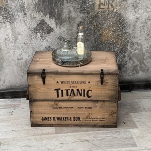 Może przedstawiać: Drewniane pudełko do przechowywania z nadrukiem "White Star Line", "R.M.S Titanic", "Southampton", "New York" i "James R. Walker & Son" na przodzie. Pudełko jest brązowe i ma metalowe zawiasy.