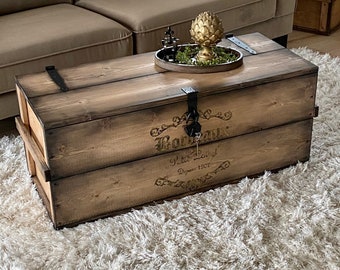 May include: A rustic wooden trunk used as a coffee table. The trunk is stained in shades of brown and features metal hardware. The text "Bordeaux" and other script is visible on the front. A decorative tray with a gold artichoke sculpture sits on top.